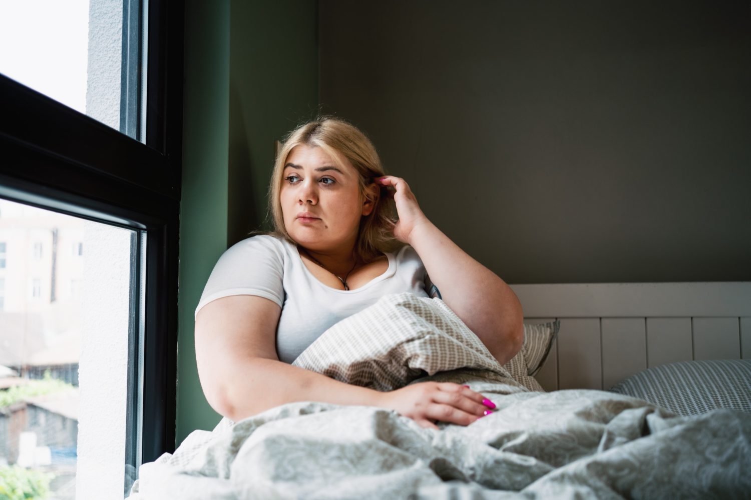 young woman sitting in her bed looking contemplatively out the window